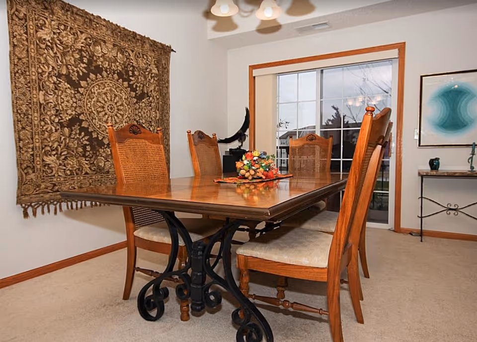 Wooden dining table with four chairs in a dining room decorated with a tapestry and sliding glass door.