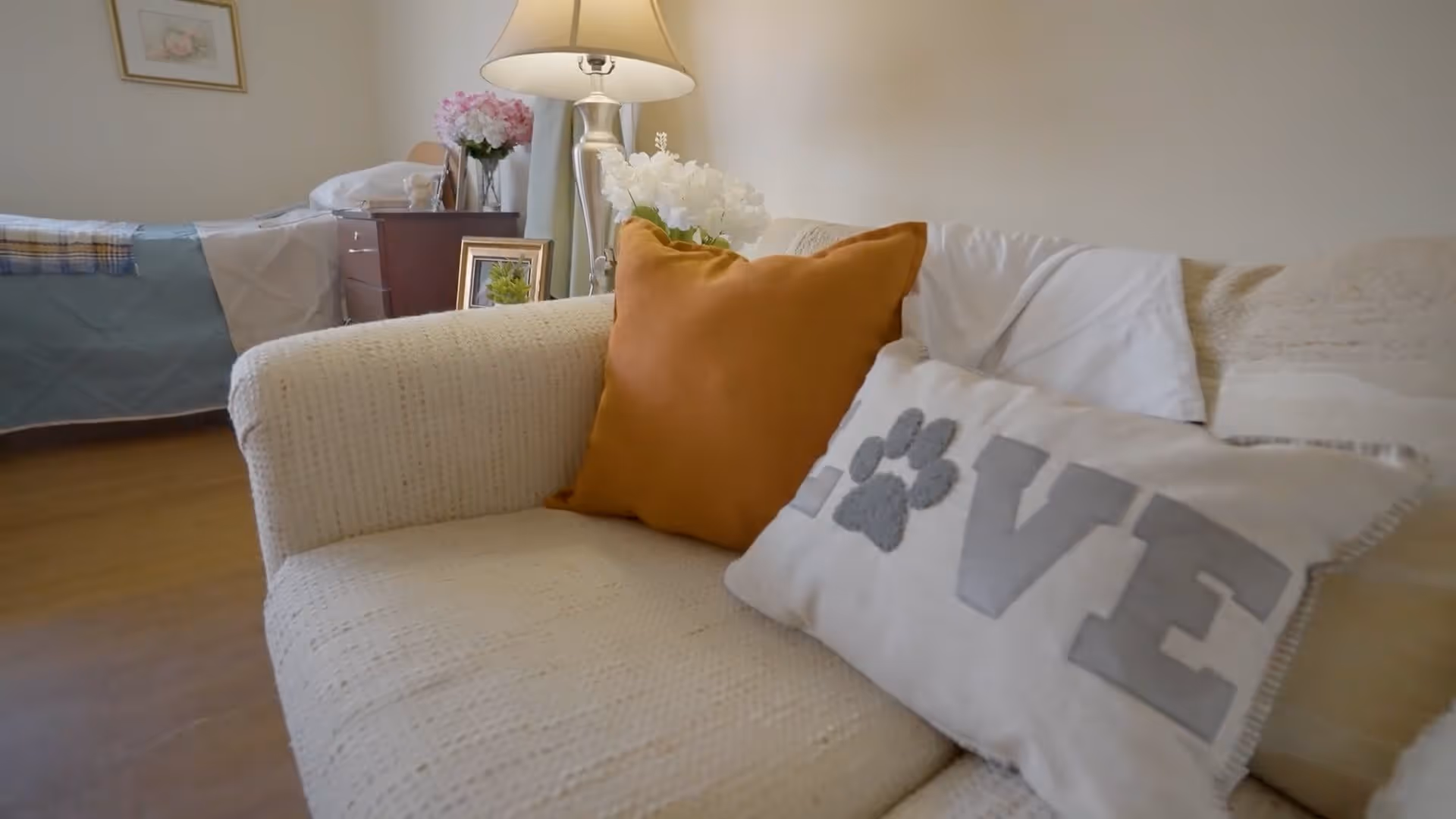 A cozy living area in an assisted living facility featuring a cream-colored textured sofa with two decorative pillows, one orange and one white with the word 'LOVE' and a paw print. In the background, there is a bed with a blue and white blanket, a wooden nightstand with a lamp, flowers, and framed pictures, all set against a light-colored wall.