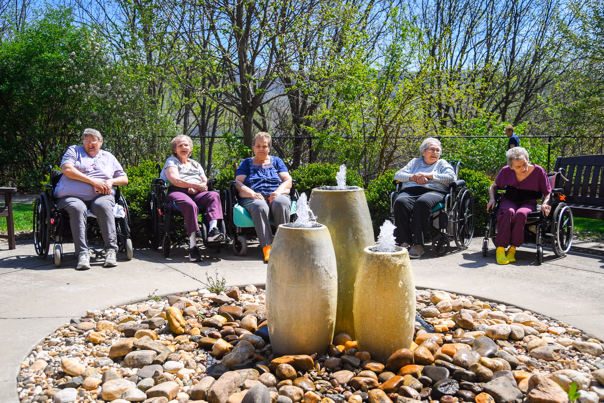 Five elderly residents in wheelchairs sit in a sunny courtyard around a three-urn stone fountain and rock garden.