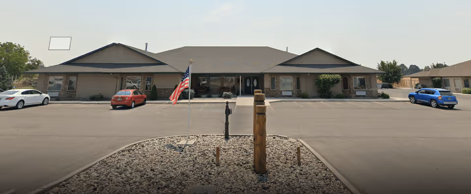 Single-story assisted living building with a central entrance, flagpole, and parking lot in front.