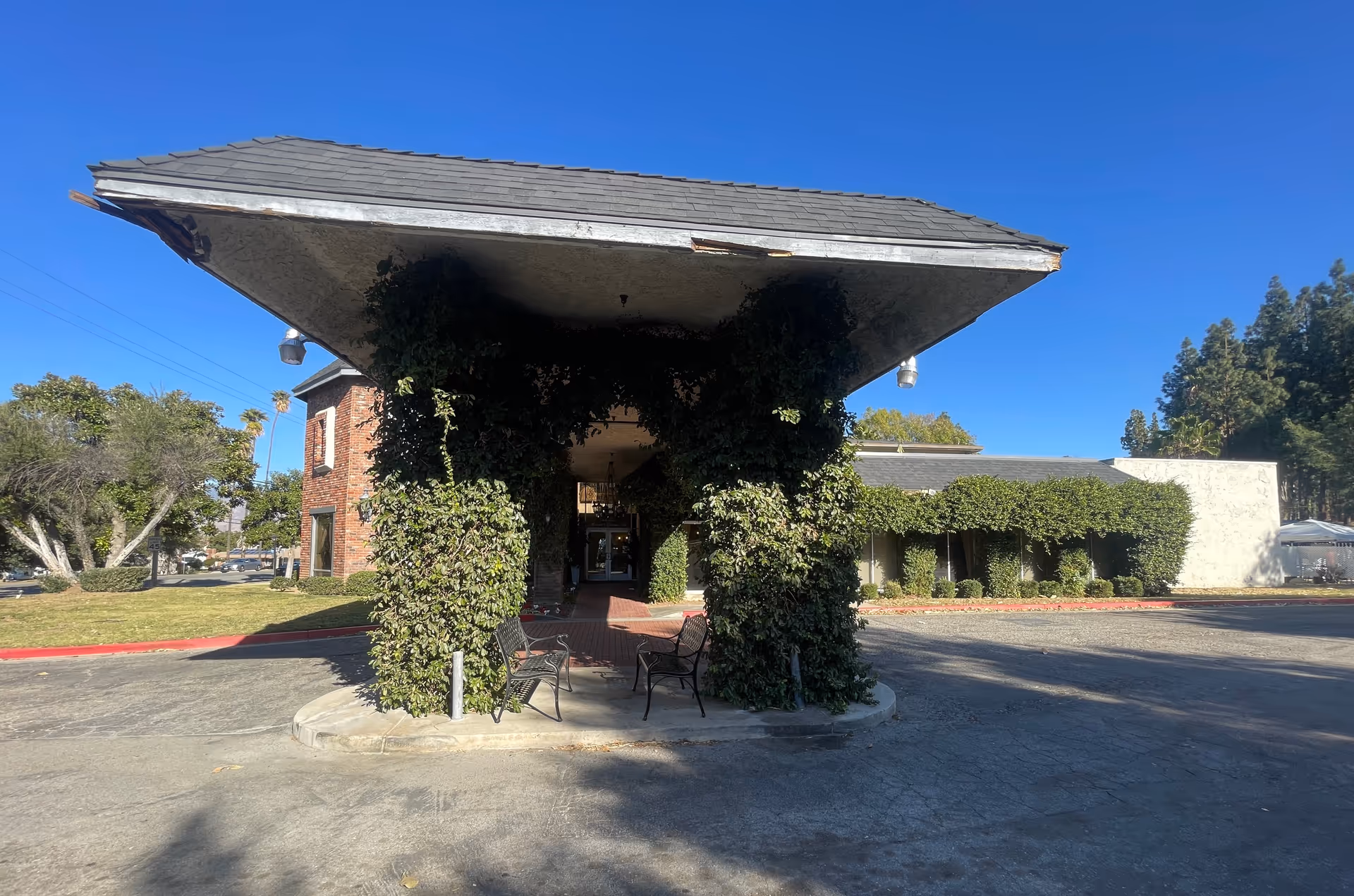 Entrance area of Braswell Family Senior Care facility with a large overhanging roof supported by pillars covered in green ivy. Two black metal benches are placed under the roof. The building exterior features brick and white walls with trimmed bushes and trees around the property under a clear blue sky.