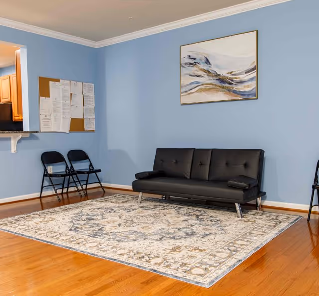 Small common room with a black futon sofa, folding chairs, a bulletin board, a framed painting on a blue wall, and a patterned area rug on hardwood floors.