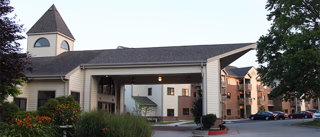 Exterior view of Unbridled Living of Columbia senior living facility showing a covered entrance with a peaked roof, surrounded by landscaped greenery and trees, with multi-story residential buildings and parked cars in the background.