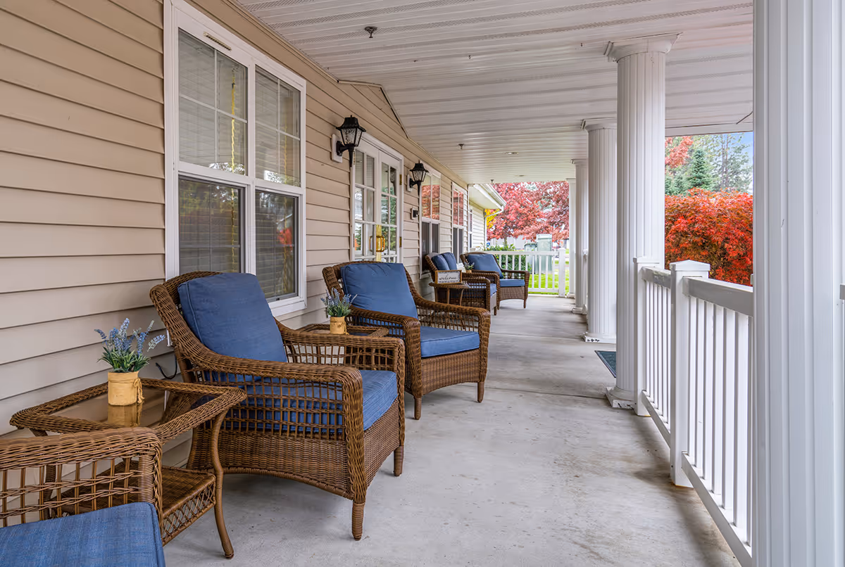 A covered outdoor porch area with several wicker chairs featuring blue cushions and small side tables with potted plants. The porch has white columns and railings, beige siding, and windows along the building wall. Trees with red and green foliage are visible in the background.