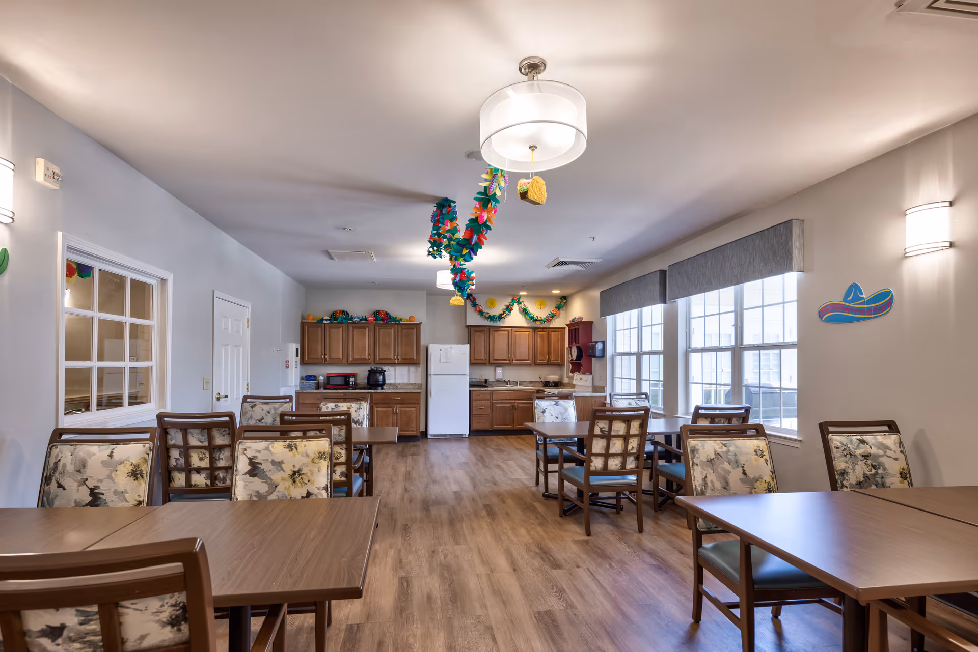 A bright and clean dining room in an assisted living facility with several wooden tables and chairs featuring floral upholstery. The room has large windows with gray valances letting in natural light. The back wall has a kitchenette area with wooden cabinets, a white refrigerator, microwave, and coffee maker. Colorful paper decorations hang from the ceiling and walls, including a sombrero on the right wall.