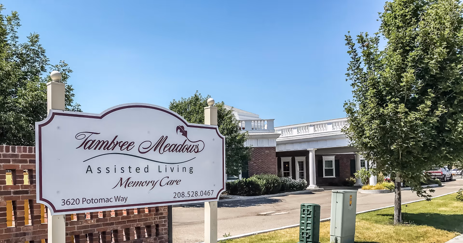 Outdoor view of Tambree Meadows Assisted Living facility with a white sign displaying the facility name, services including memory care, address, and phone number. The building is partially visible in the background with a brick exterior and white columns under a clear blue sky.