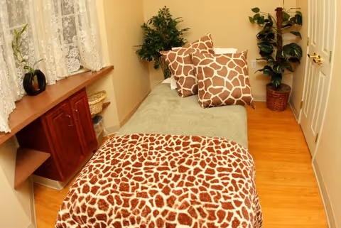 A small bedroom with a single bed covered in a green blanket and a giraffe-patterned throw and pillows. The room has wooden flooring, a wooden cabinet with lace curtains above it, and two potted plants in the corners.