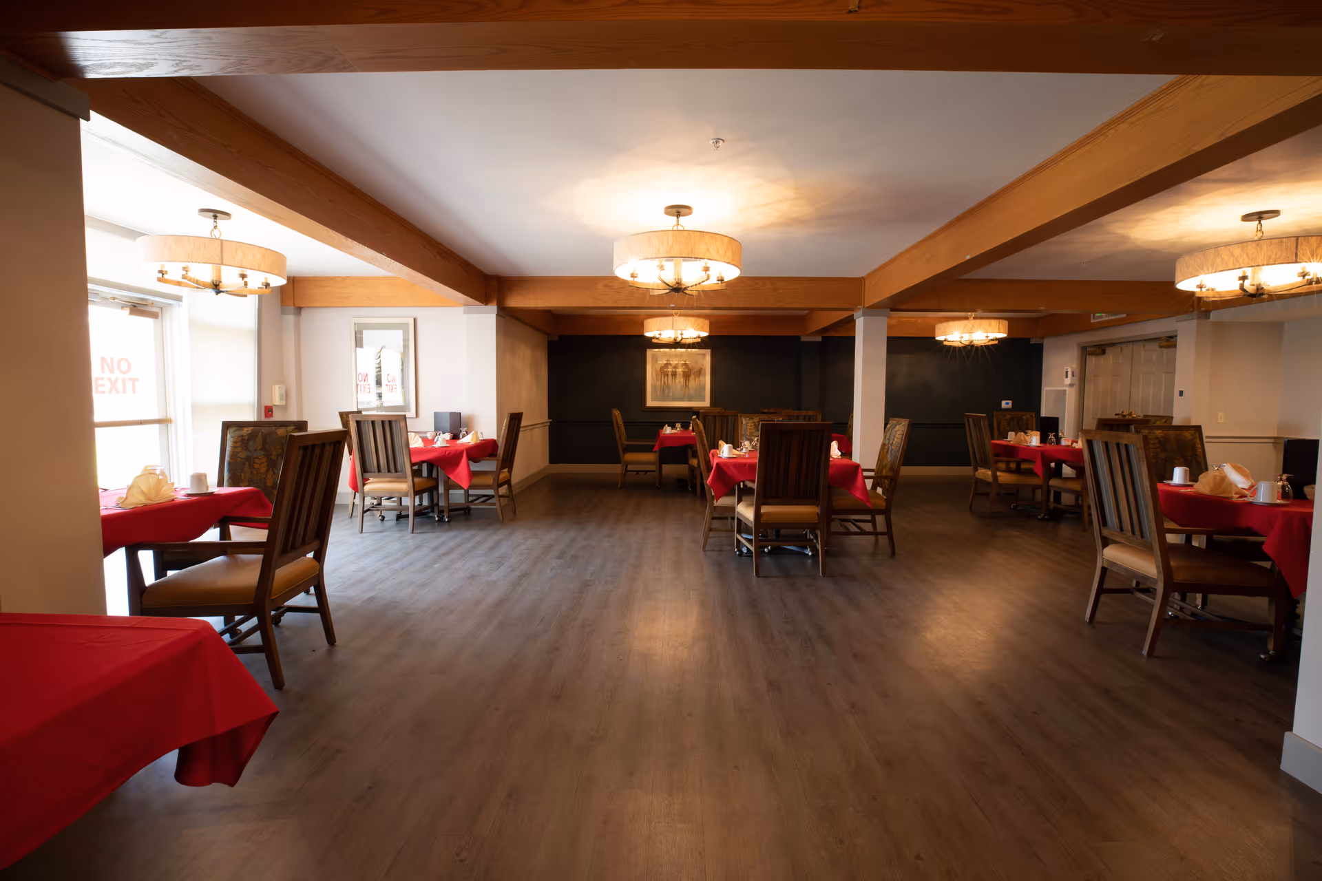 A dining room with several tables covered in red tablecloths, each set with napkins, cups, and plates. The room features wooden beams on the ceiling, modern chandeliers, and wooden chairs with cushioned seats. The floor is a wood-like material, and there is a 'No Exit' sign visible on a glass door to the left.