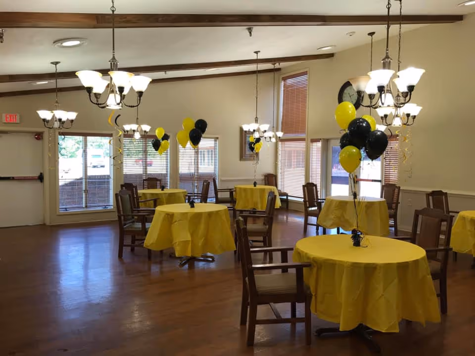 A spacious dining room with several round tables covered in yellow tablecloths. Each table has a centerpiece of black and yellow balloons. Wooden chairs surround the tables, and multiple chandeliers hang from the ceiling providing warm lighting. Large windows with blinds allow natural light to enter the room.