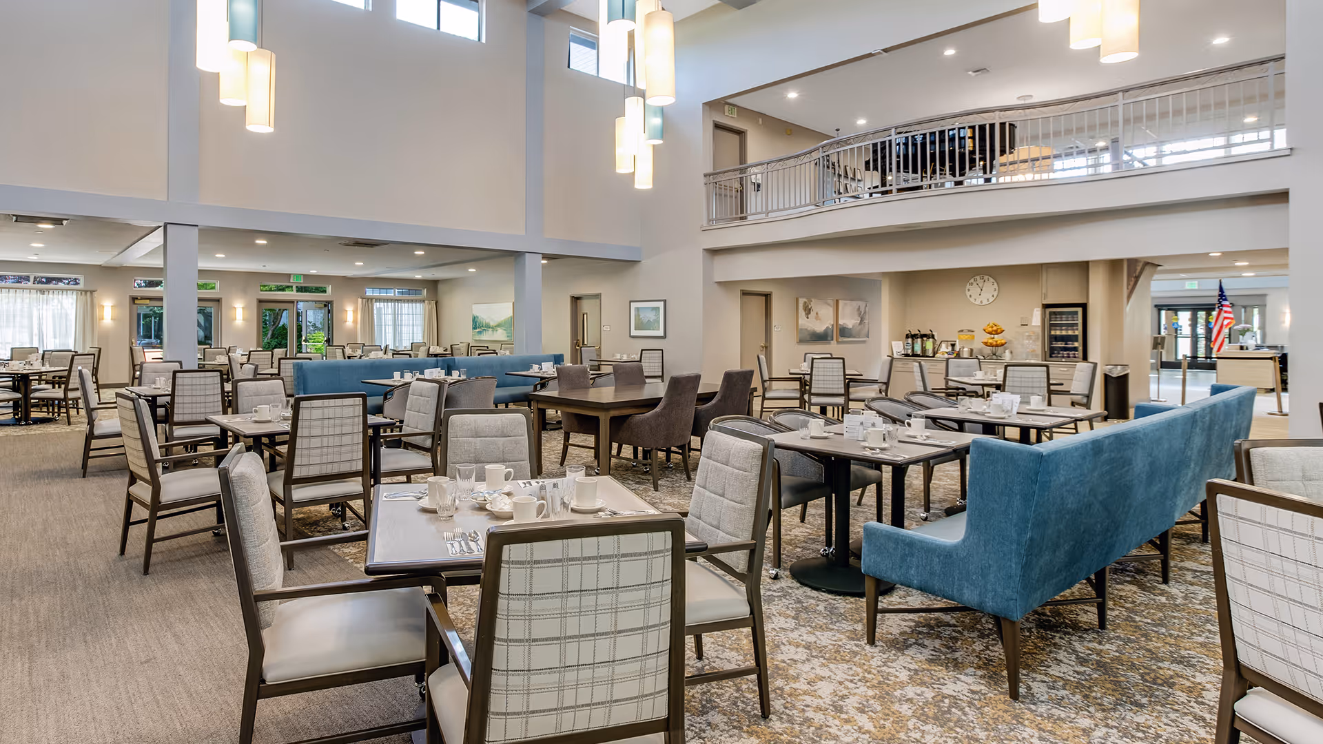 A spacious and well-lit dining room in a senior living facility with multiple tables and chairs arranged neatly. The room features high ceilings with hanging pendant lights, large windows allowing natural light, and a mix of cushioned chairs and blue upholstered bench seating. The walls are decorated with framed artwork, and there is a beverage station with coffee urns and fruit in the background. An American flag is visible near the entrance.