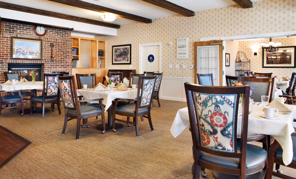 A cozy dining room with several tables covered in white tablecloths, each set with napkins, cups, and small flower arrangements. The room features patterned wallpaper, a brick fireplace with a clock and framed artwork above it, and wooden chairs with colorful patterned backs. There are wooden beams on the ceiling and a carpeted floor.