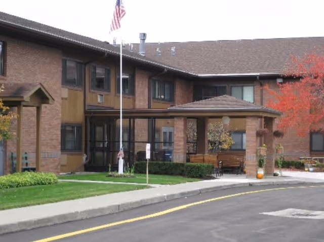 Front entrance of a two-story brick senior living facility with a covered porte-cochere, flagpole, benches, and autumn landscaping.