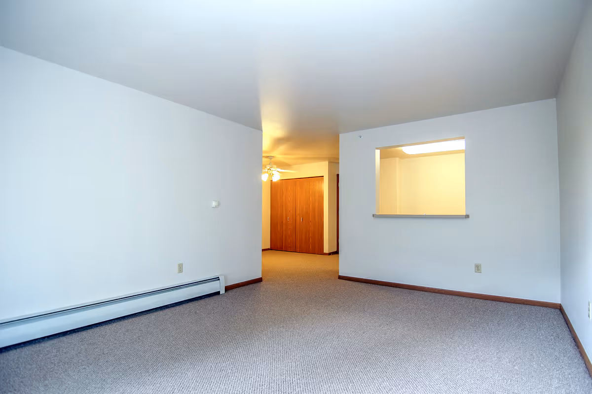 Empty room with beige carpet and white walls, featuring a small pass-through window to another room, a ceiling fan with lights, and wooden closet doors in the background.