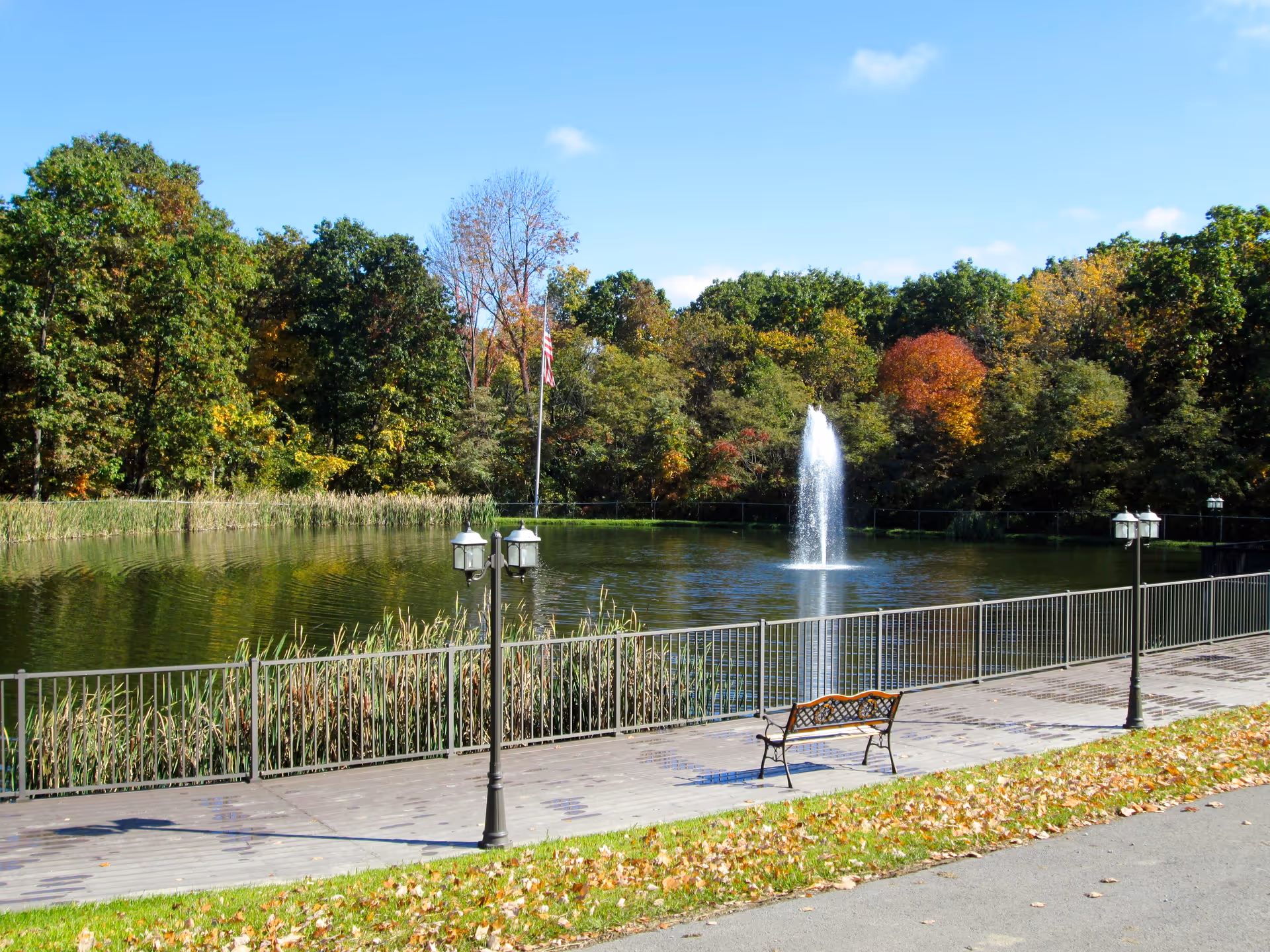 A peaceful outdoor scene featuring a pond with a water fountain in the center, surrounded by trees with autumn foliage. There is a paved walkway with a metal railing along the pond, a bench, and vintage-style street lamps. The sky is clear and blue.