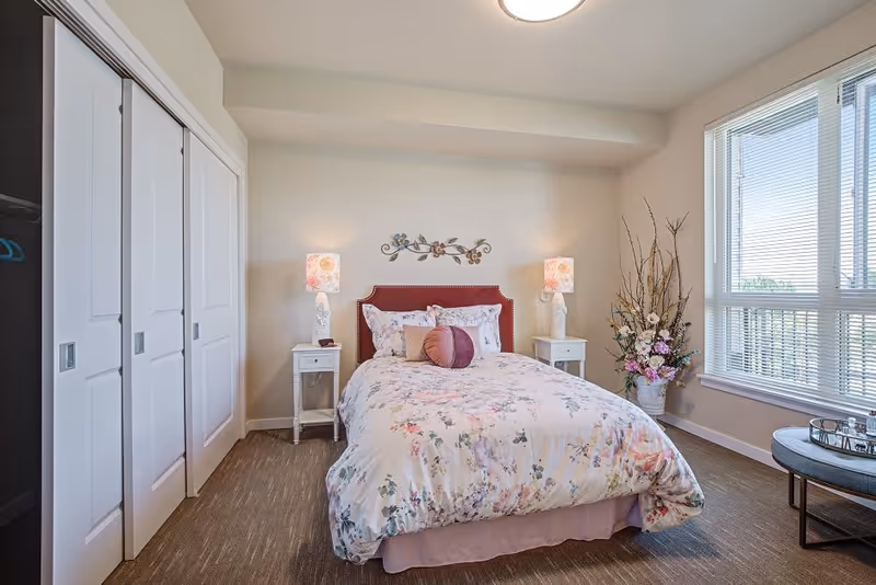 A bright and cozy bedroom featuring a bed with floral bedding and decorative pillows, flanked by two white nightstands each with a floral lamp. There is a large window with blinds on the right side, a decorative floral arrangement in a vase near the window, and a small round table with a tray. On the left side, there are white sliding closet doors.