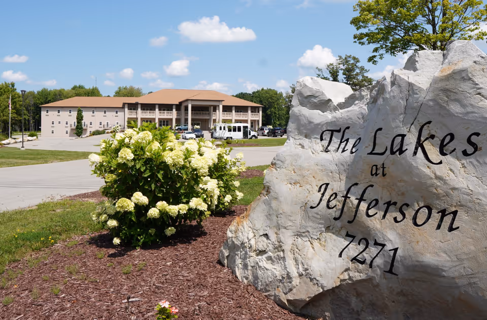 Front exterior of The Lakes at Jefferson senior living building with a large engraved stone sign and flowering shrubs.