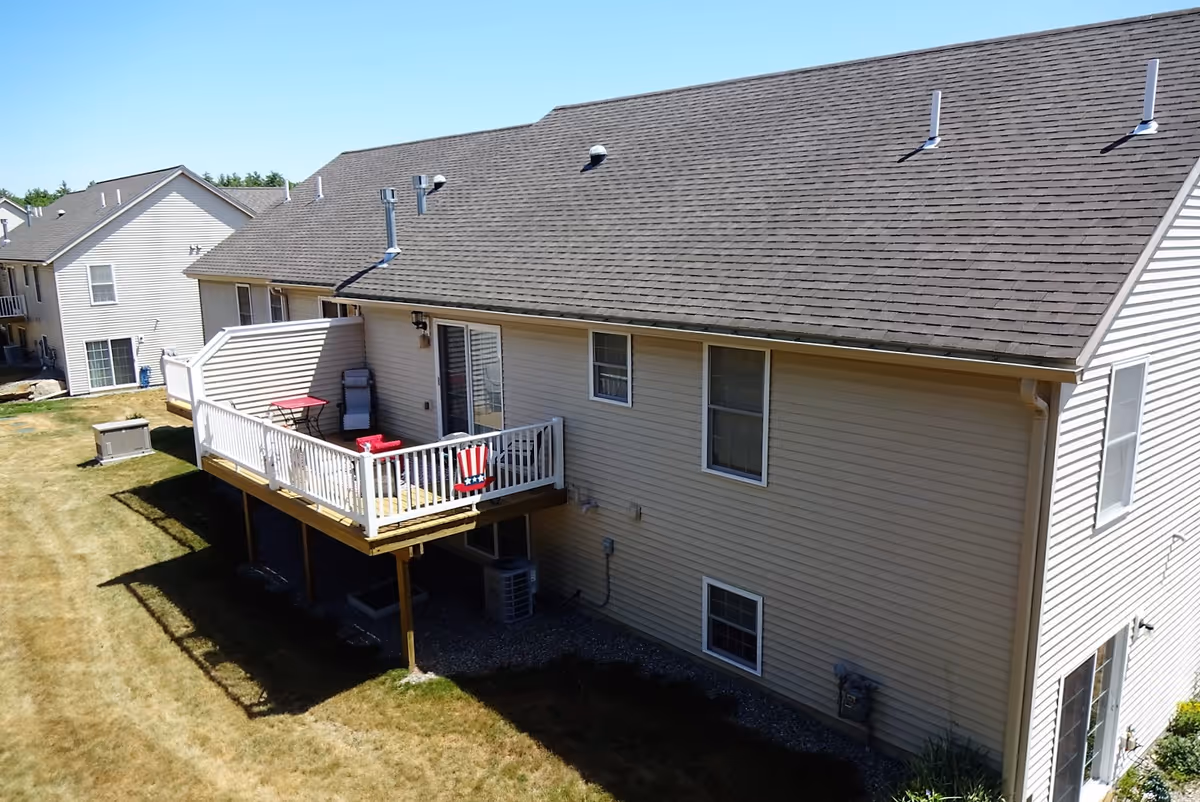 Exterior view of a beige two-story residential building with a wooden deck featuring white railings, a red chair, and a small table. The building has multiple windows and a shingled roof. The surrounding area includes a grassy yard and neighboring similar buildings.