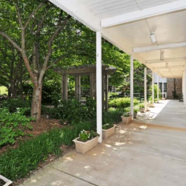 Covered walkway with white pillars and ceiling lights alongside a garden area with green trees, shrubs, and a wooden pergola in the background.
