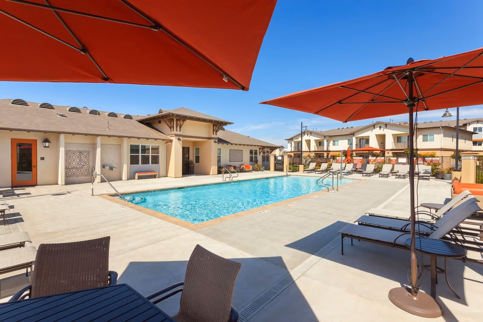 Outdoor swimming pool area at Villa del Sol facility with lounge chairs, tables, and large red umbrellas under a clear blue sky. Surrounding buildings and a poolside seating area are visible.