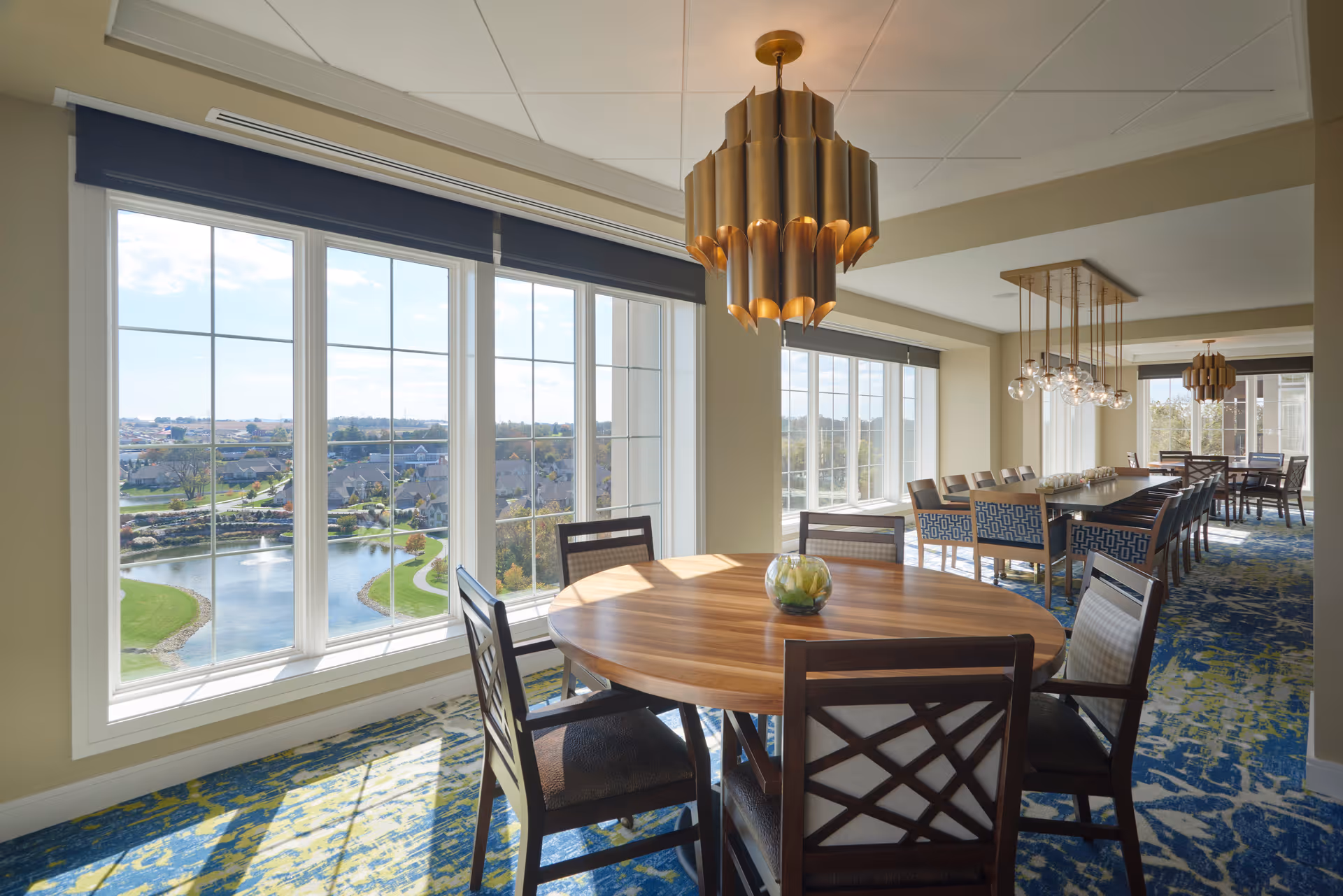 A bright dining room with large windows offering a view of a pond and houses outside. The room features a round wooden table with four chairs in the foreground and a long rectangular table with multiple chairs in the background. Two modern pendant lights hang from the ceiling, and the floor is covered with a blue and green patterned carpet.