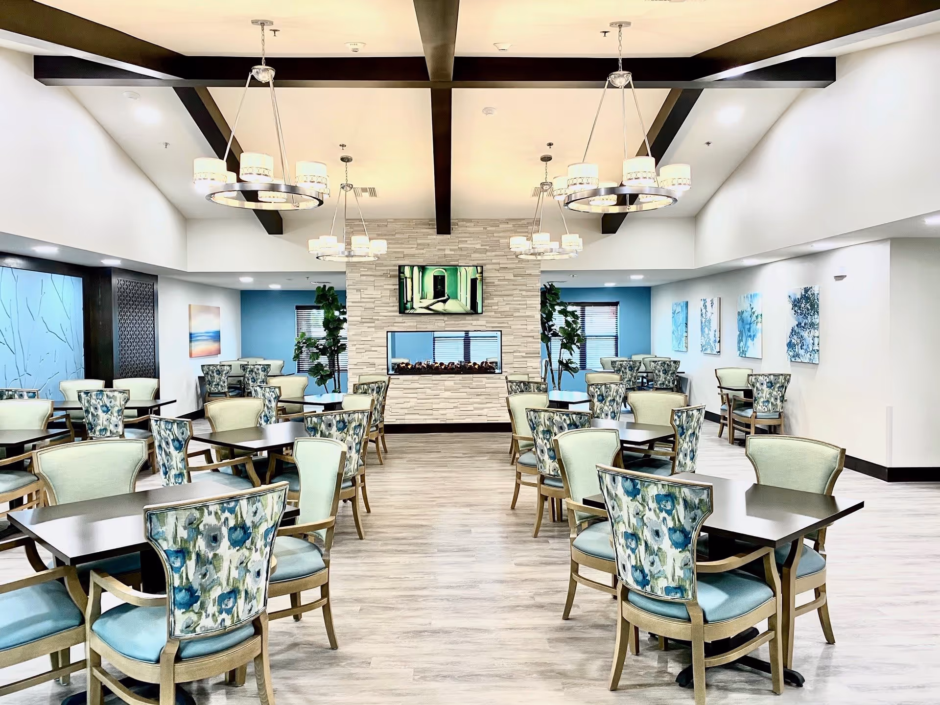 A bright and spacious dining room with multiple tables and chairs arranged neatly. The chairs have a floral pattern on the backrest and light blue cushions. The room features a modern fireplace with a TV mounted above it, decorative ceiling beams, and elegant circular chandeliers. The walls are decorated with blue and white artwork and plants are placed near the windows.