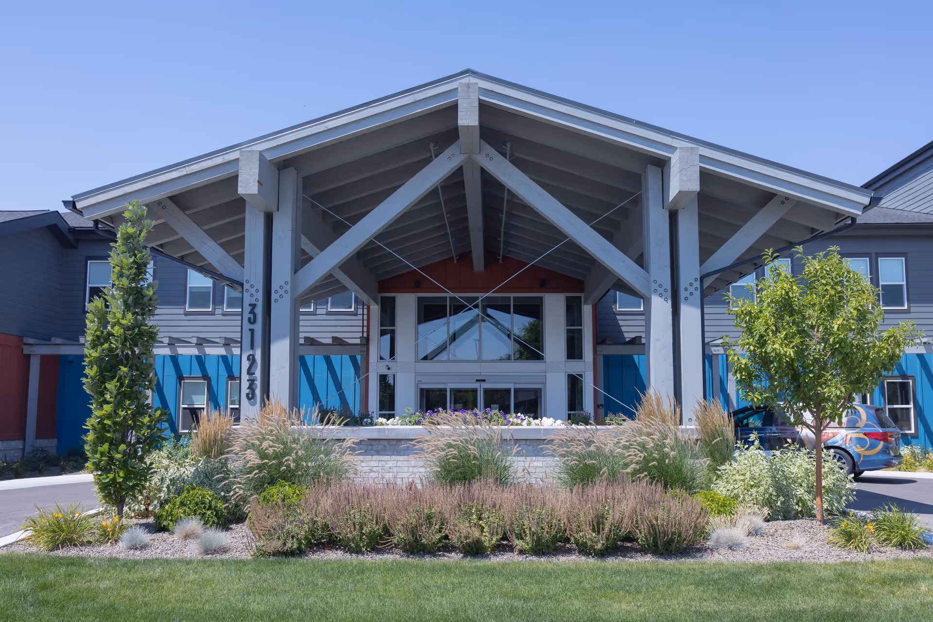 Covered front entrance of a modern building with a large wooden porte-cochere, landscaped plantings, and a glass entry.