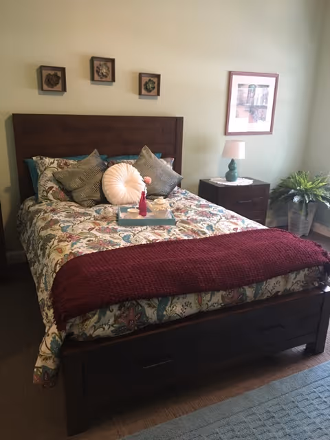 A neatly made bed with a floral patterned bedspread, multiple pillows including a round white one, and a maroon throw blanket at the foot. A wooden headboard and footboard frame the bed. On the bed is a tray with a teapot, cup, and a small vase with a flower. To the right of the bed is a wooden nightstand with a green lamp and a framed picture hanging above it. A potted plant is placed on the floor near the nightstand. The room has light-colored walls and carpeted flooring.
