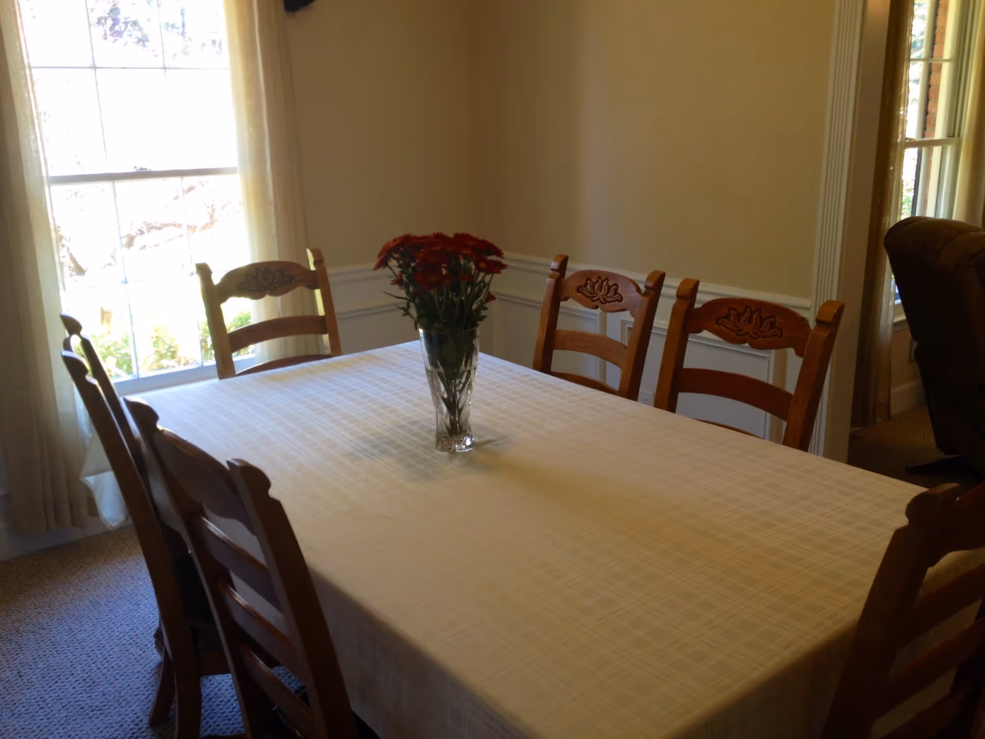 A dining room with a rectangular table covered with a white tablecloth. There are six wooden chairs around the table, and a vase with red flowers is placed in the center. The room has beige walls, a window with sheer curtains letting in natural light, and a carpeted floor.