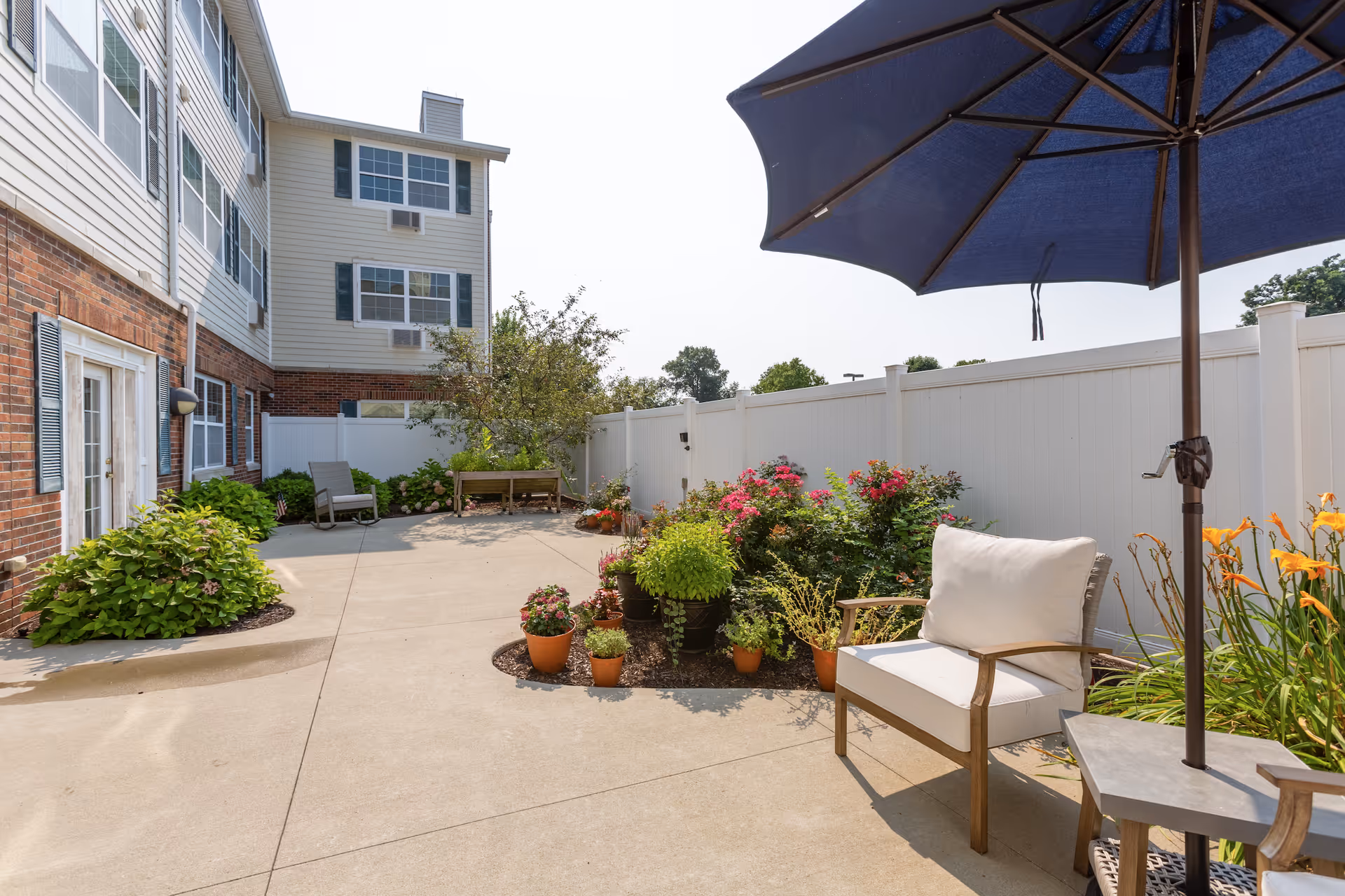 Outdoor patio area at StoryPoint Granger with a concrete floor, potted plants, flowers, a white privacy fence, a cushioned chair under a large blue umbrella, and a bench near the building. The building exterior features brick and siding with multiple windows.