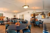 A spacious dining room with multiple round tables and green cushioned chairs arranged neatly. Each table has a small floral centerpiece. The room has wooden flooring, white walls, and ceiling lights. In the background, there is a kitchen area with a serving counter and cabinets.
