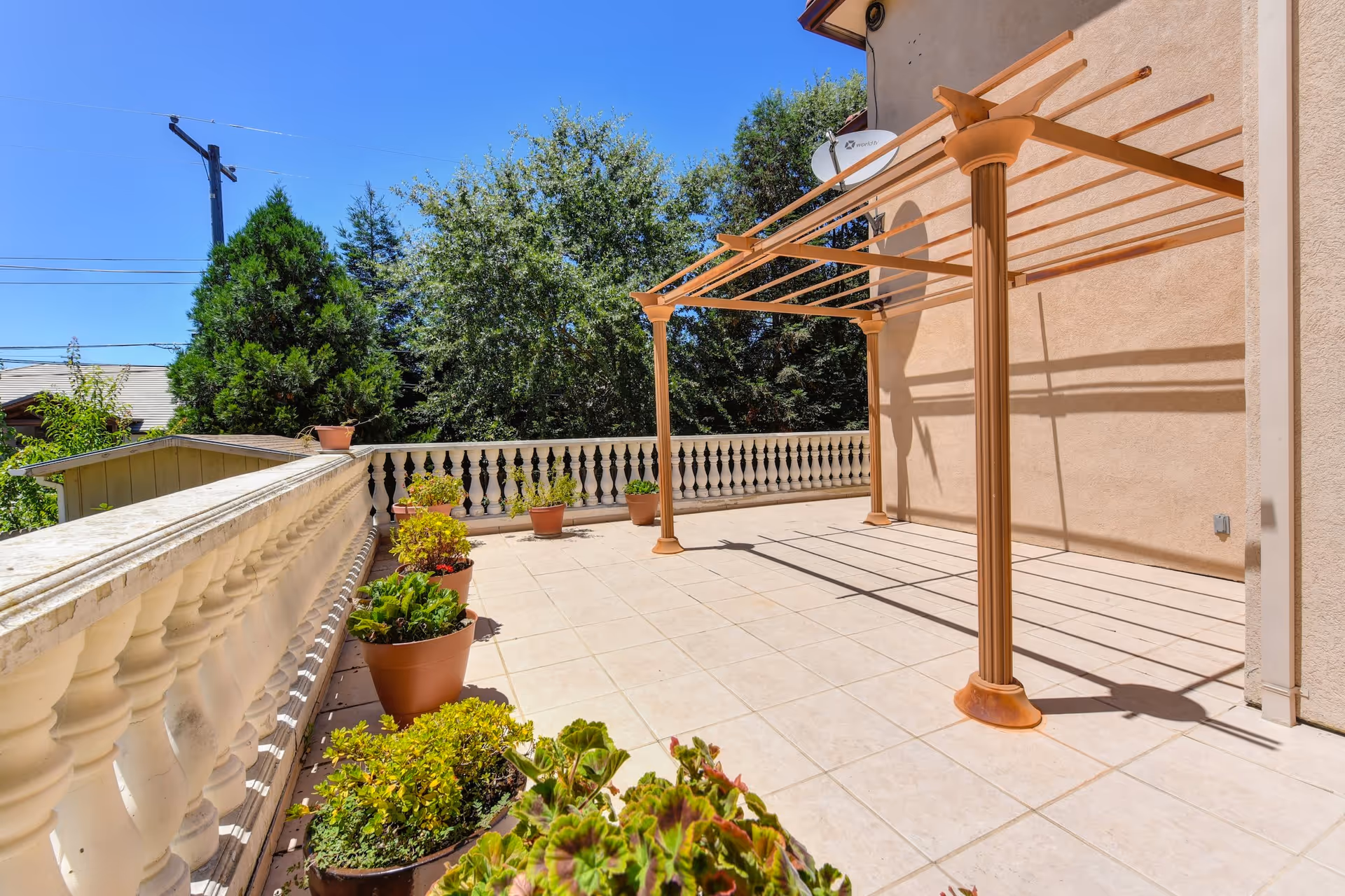 A spacious outdoor patio area with tiled flooring, a beige pergola structure attached to the building, and several potted plants lined along a decorative white balustrade. Trees and a clear blue sky are visible in the background.