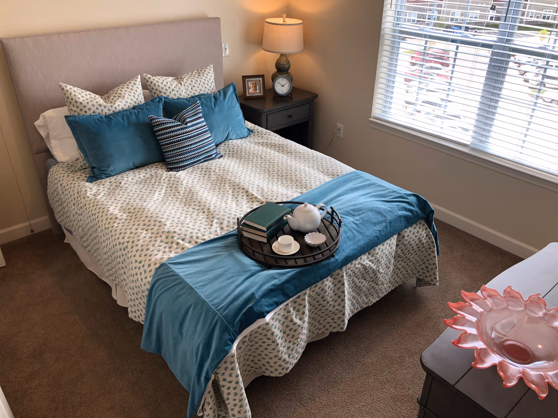 A neatly made bed with patterned white and teal bedding and multiple pillows in a bedroom. On the bed is a black tray holding a white teapot, a cup and saucer, a small dish, and two green books. Next to the bed is a nightstand with a lamp, a clock, and a framed photo. A window with white blinds lets in natural light, and a dresser with a decorative pink glass bowl is visible in the foreground.