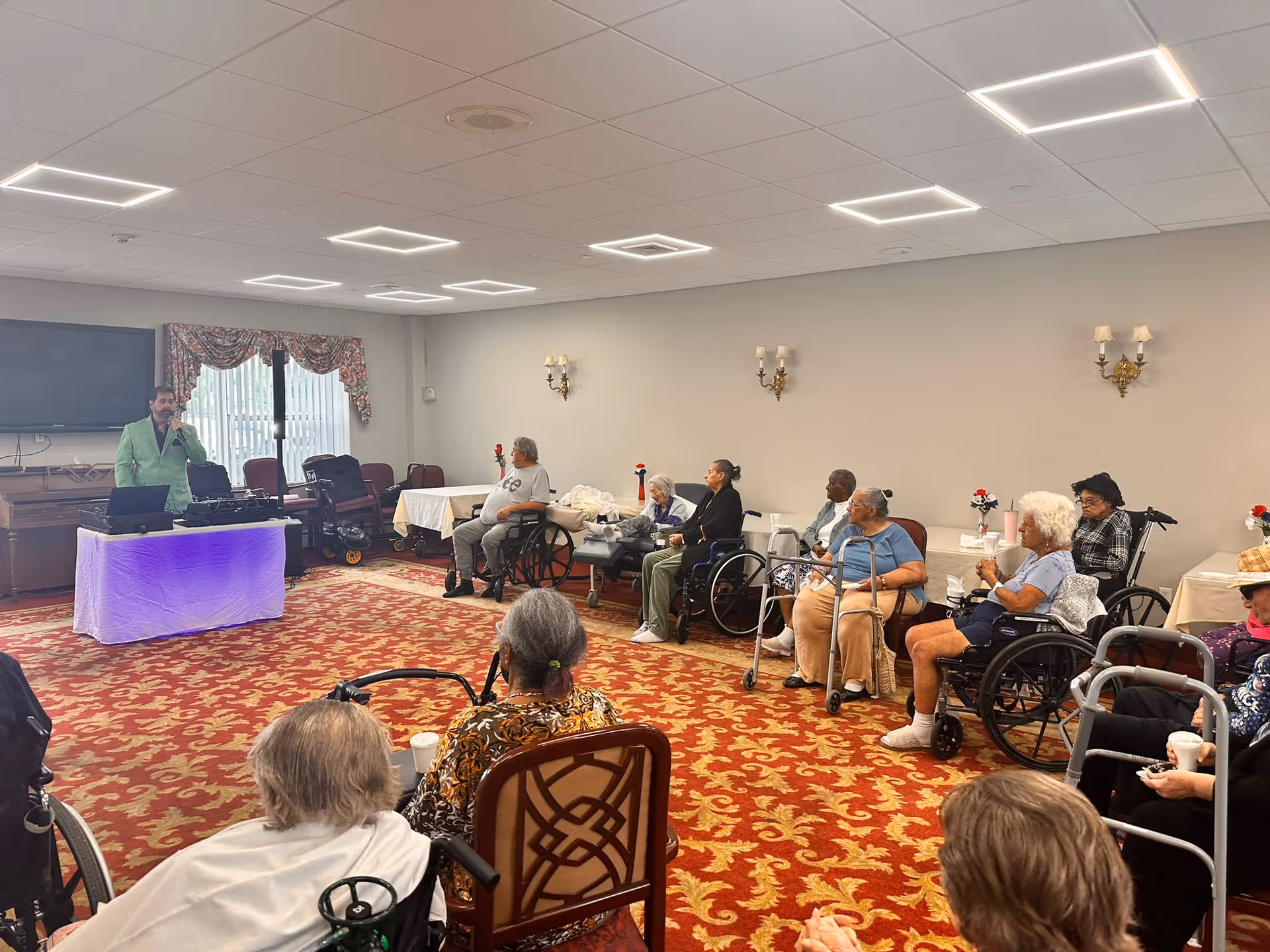 A group of elderly people, many in wheelchairs, seated in a room with a red and gold patterned carpet. They are facing a man in a green jacket standing near a table with electronic equipment and a purple light underneath. The room has beige walls with wall sconces and a window with floral curtains.