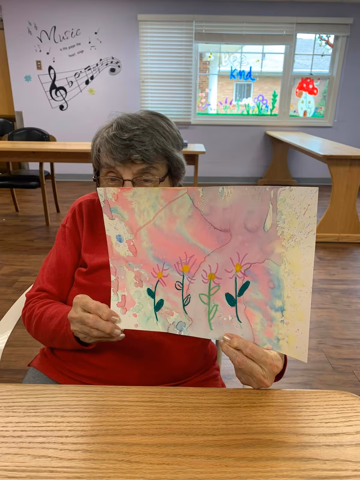 An elderly woman wearing glasses and a red sweater is sitting at a wooden table in a room. She is holding up a colorful painting of four flowers with pink, yellow, and green colors. Behind her, there is a wall with musical notes and the word 'Music' written on it, and a window decorated with drawings and the words 'Be kind'. The room has wooden floors and tables with chairs.