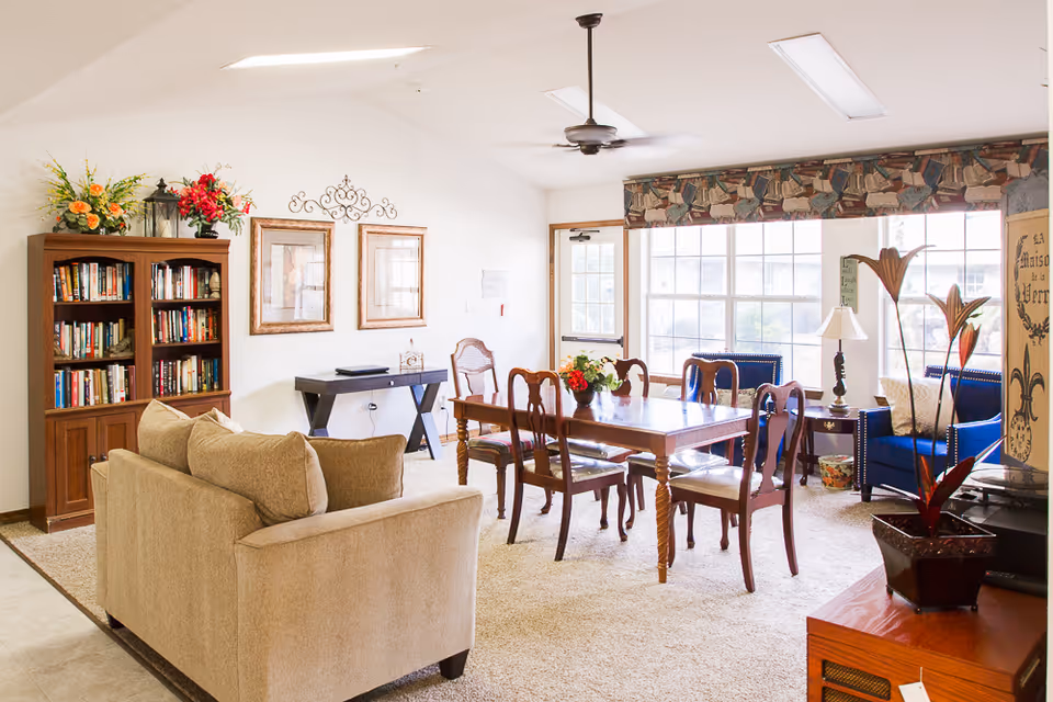 A bright and cozy living room and dining area in Berryhill Manor featuring a beige sofa, a wooden dining table with six chairs, a bookshelf filled with books, decorative flowers, framed artwork on the wall, a ceiling fan, large windows with patterned valances, and blue armchairs near a side table with a lamp.
