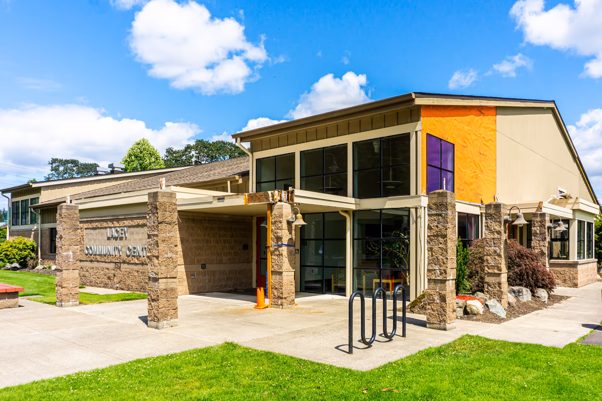 Exterior view of the Lacey Community Center building on a sunny day with blue sky and scattered clouds. The building features large windows, stone pillars, and a small covered entrance area. There is a bike rack and some landscaping with grass and bushes around the building.