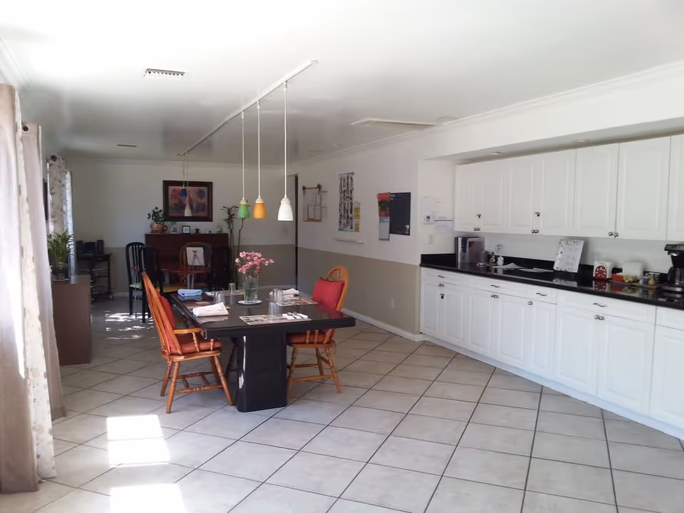 A bright dining area with a black rectangular table set for four with napkins, glasses, and a vase of pink flowers. Four wooden chairs with red cushions surround the table. To the right is a long white cabinet with black countertop holding a coffee maker and other kitchen items. The floor is tiled, and the walls are white with a beige lower half. Pendant lights hang above the table. In the background, there is another small table with chairs and framed artwork on the wall.