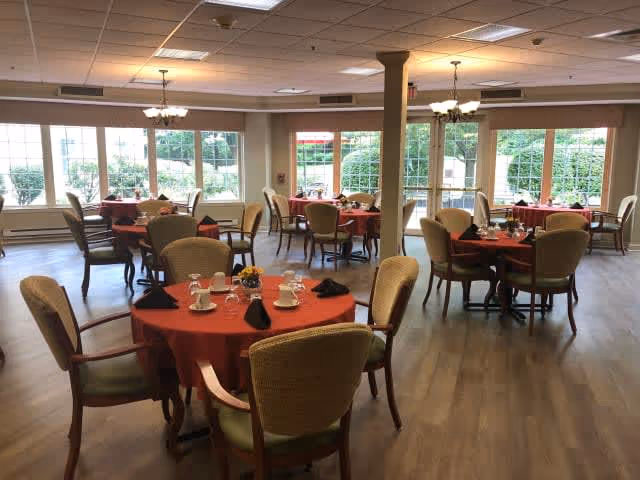 A dining room with several round tables covered with red tablecloths, each set with cups, glasses, and black napkins. The room has large windows letting in natural light and wooden flooring. Comfortable chairs surround each table, and chandeliers hang from the ceiling.