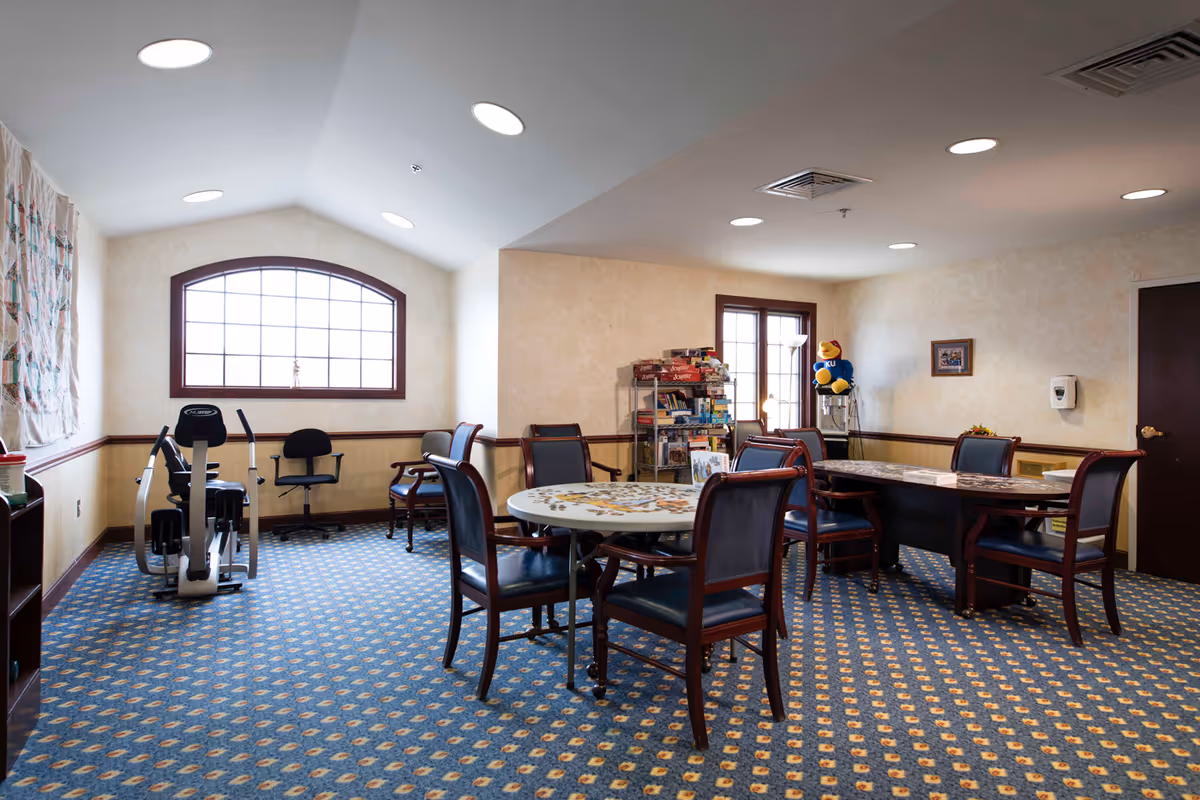 A well-lit activity room with patterned blue carpet and beige walls. The room contains two tables with chairs around them, one round and one rectangular, both with puzzles in progress. There is a rack filled with board games and books near a window with wooden trim. An exercise bike and an office chair are positioned near a large arched window. A plush bird wearing a KU shirt sits on a water dispenser in the corner. The ceiling has recessed lighting and an air vent.