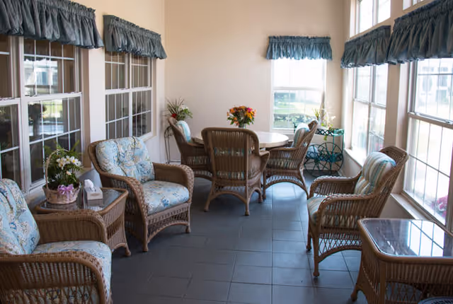A sunlit room with multiple wicker chairs and a round table in the center. The chairs have floral and striped cushions. The room has large windows with blue valances, letting in natural light. There are small tables with flower arrangements and a decorative plant stand near the windows.