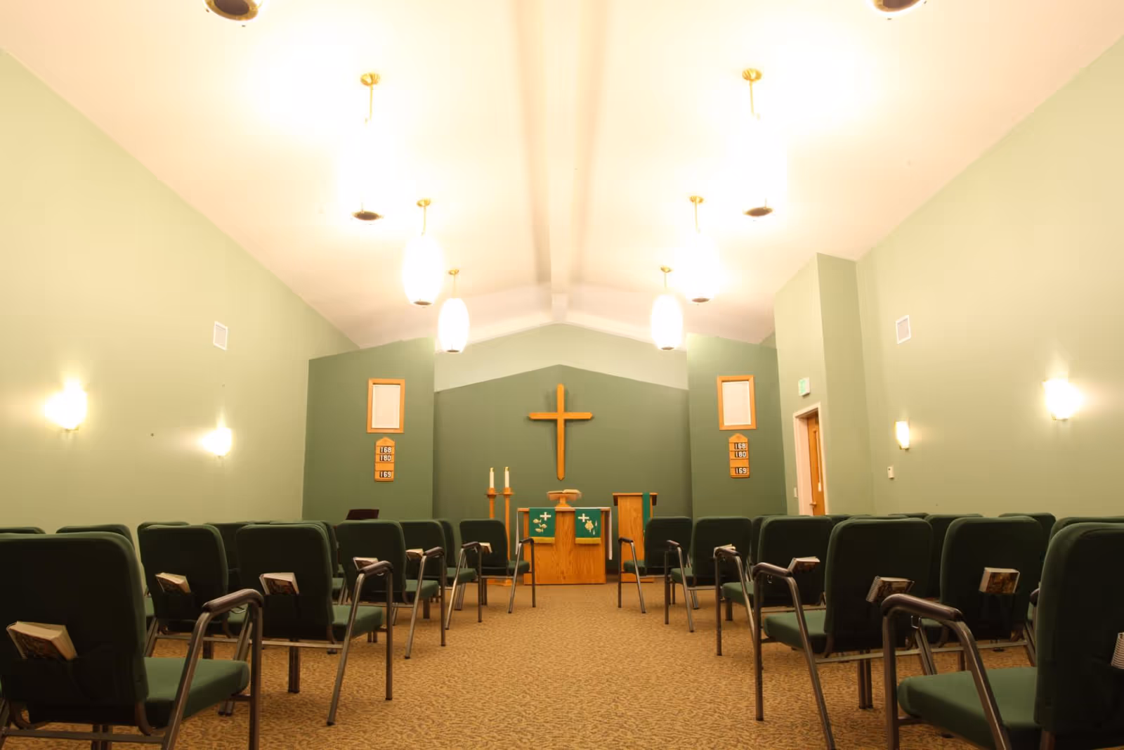 Interior view of a chapel with green walls, rows of green cushioned chairs with hymnals, a wooden altar with a green cloth, two candles, and a wooden cross mounted on the wall behind the altar. The ceiling has several hanging light fixtures.