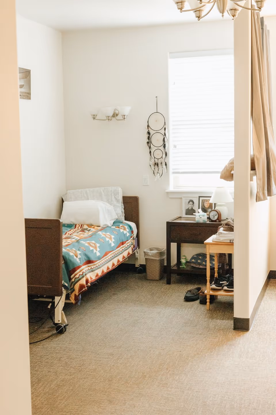 A small bedroom with a single bed covered with a colorful patterned blanket. There is a pillow and a folded blanket on the bed. Next to the bed is a dark wooden nightstand with framed photos, a lamp, an alarm clock, and some tissues. A dreamcatcher hangs on the wall beside a window with closed blinds. A small trash can is on the floor near the nightstand, and a wooden shoe rack with shoes is partially visible. The room has beige walls and carpeted flooring.