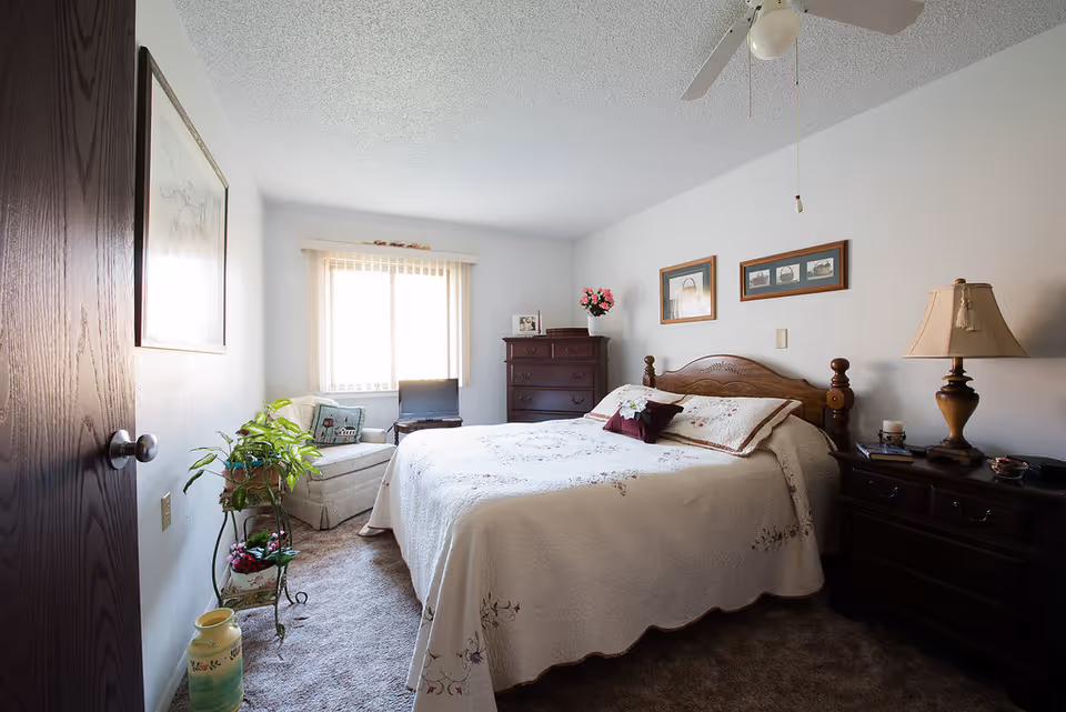 A cozy bedroom with a large bed covered in a white floral quilt. There is a wooden headboard and matching dark wood nightstands on either side of the bed, one with a lamp and the other with a candle and small items. A wooden dresser with a vase of pink flowers and framed photos sits against the far wall. A cushioned armchair with a decorative pillow is placed near a window with vertical blinds, and a small TV is on a stand next to the chair. A green potted plant is on a metal plant stand near the door, and a framed picture hangs on the wall to the left.