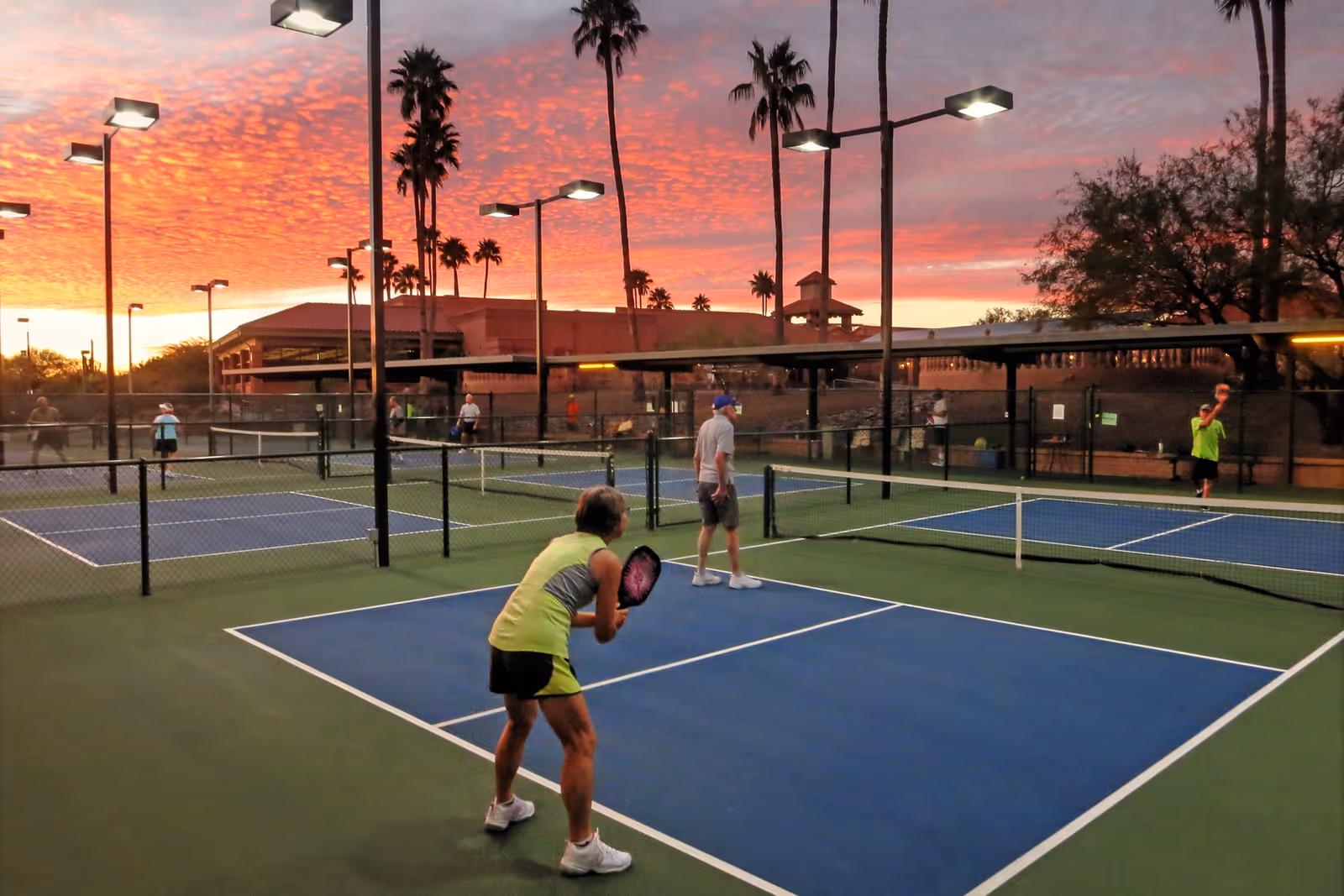 People playing pickleball on outdoor courts at sunset with a vibrant orange and pink sky, surrounded by palm trees and lit by tall floodlights.