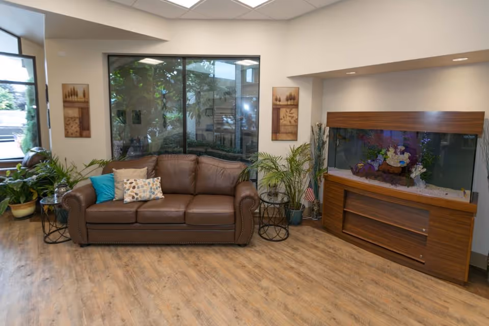 A cozy living room area with a brown leather sofa adorned with three decorative pillows. The room features large windows with a view of greenery outside, two framed artworks on the wall, several potted plants, and a large wooden aquarium filled with colorful fish and decorations. The floor is wooden, and the space is well-lit with ceiling lights.