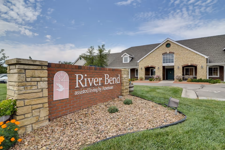 Exterior view of the River Bend assisted living facility showing a brick and stone sign with the facility name and a large building with stone and brick facade in the background under a partly cloudy sky.