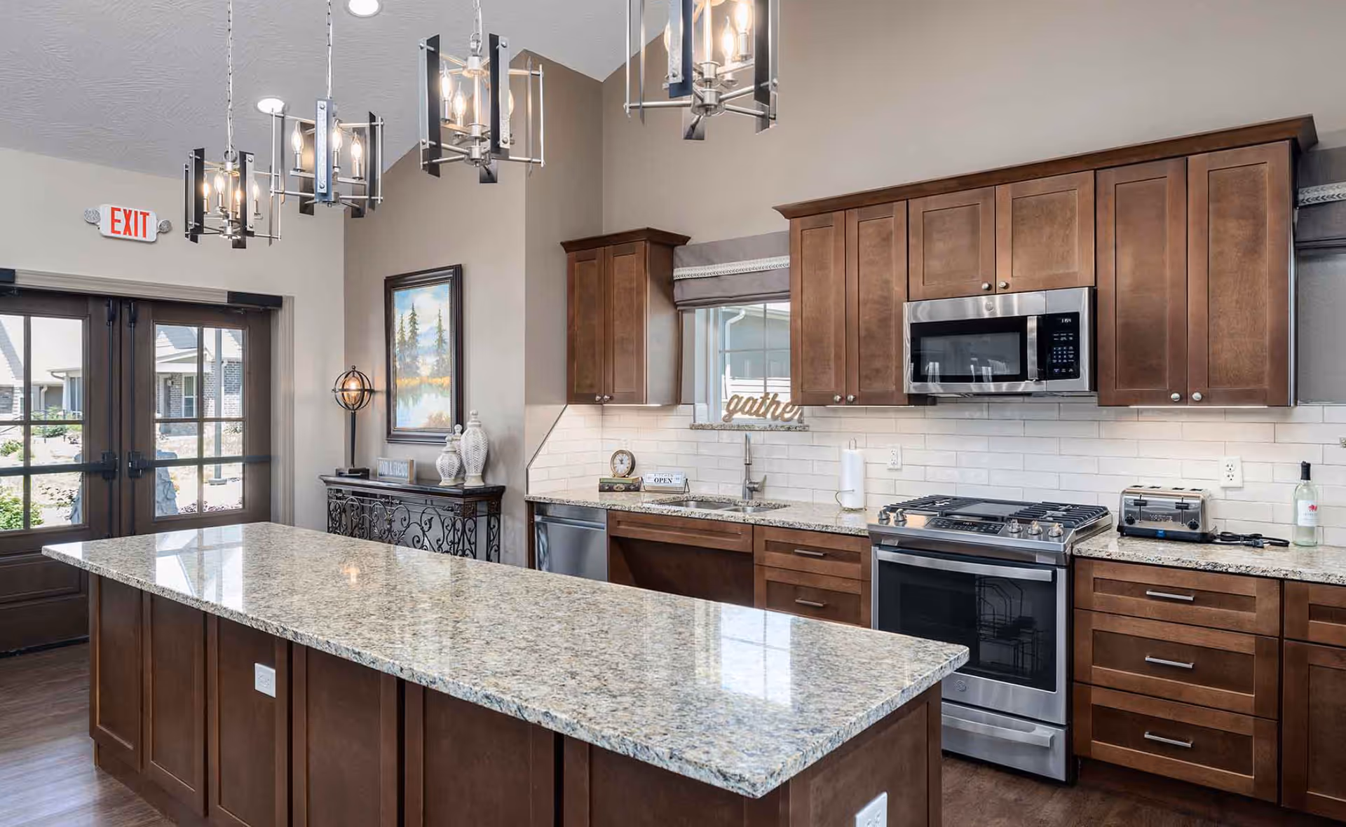 Bright communal kitchen featuring a long granite island, pendant lighting, stainless steel appliances and dark wood cabinets.