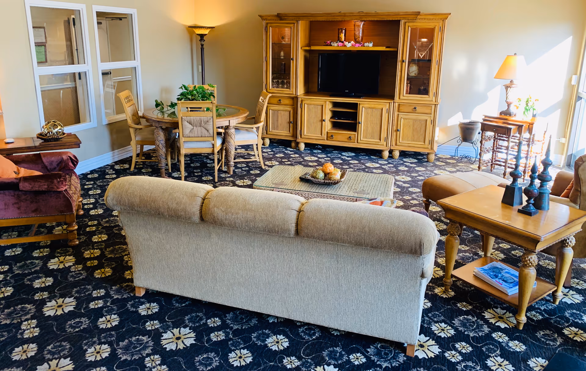 A cozy living room area with a beige sofa facing a wooden entertainment center with a TV. There is a round wooden dining table with four chairs and a potted plant on top. The room has patterned carpet flooring and is well-lit with natural light coming from a window on the right. Additional furniture includes a side table with decorative items and a lamp, a purple armchair, and a tall floor lamp in the corner.