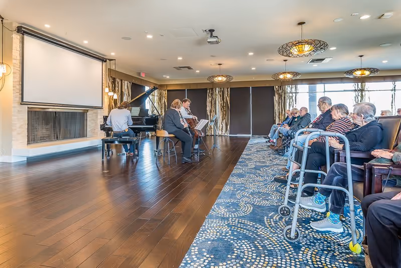 A group of elderly people seated in chairs along the right side of a room, attentively watching a small musical performance. Three musicians are playing instruments including a piano and a cello near a fireplace and a large projection screen. The room has wooden floors, patterned carpet, and decorative ceiling lights.