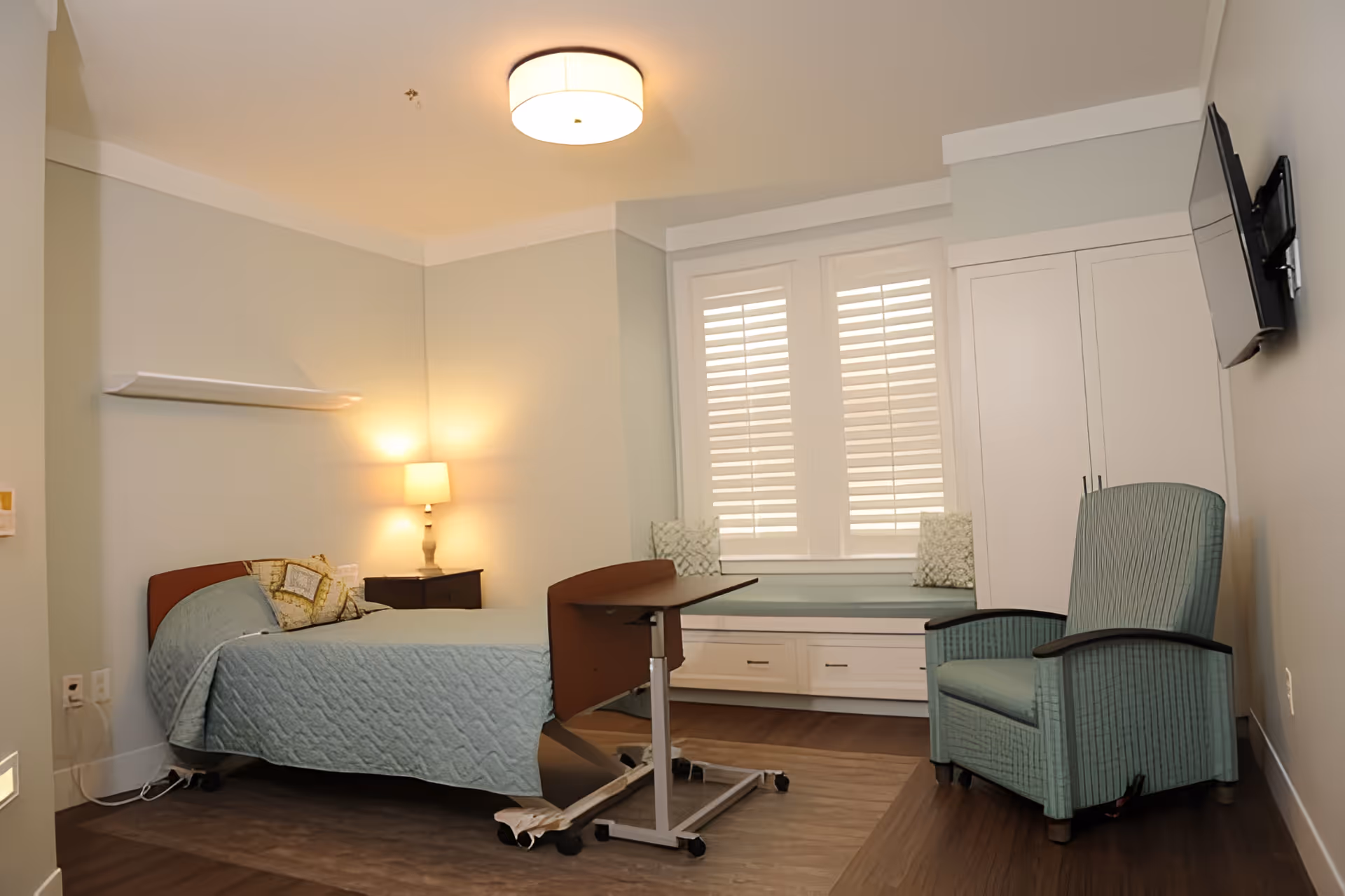 A neatly arranged bedroom in a senior living facility with a single bed covered in a light blue quilt, a bedside table with a lamp, a movable overbed table, a cushioned window seat with pillows, a green armchair, and a wall-mounted TV. The room has light-colored walls and wooden flooring.